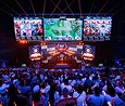 Een drukbezochte esports-arena met een intense sfeer; op grote schermen wordt een multiplayergame vertoond. Een enthousiast publiek kijkt toe, met lichtstokken in de hand